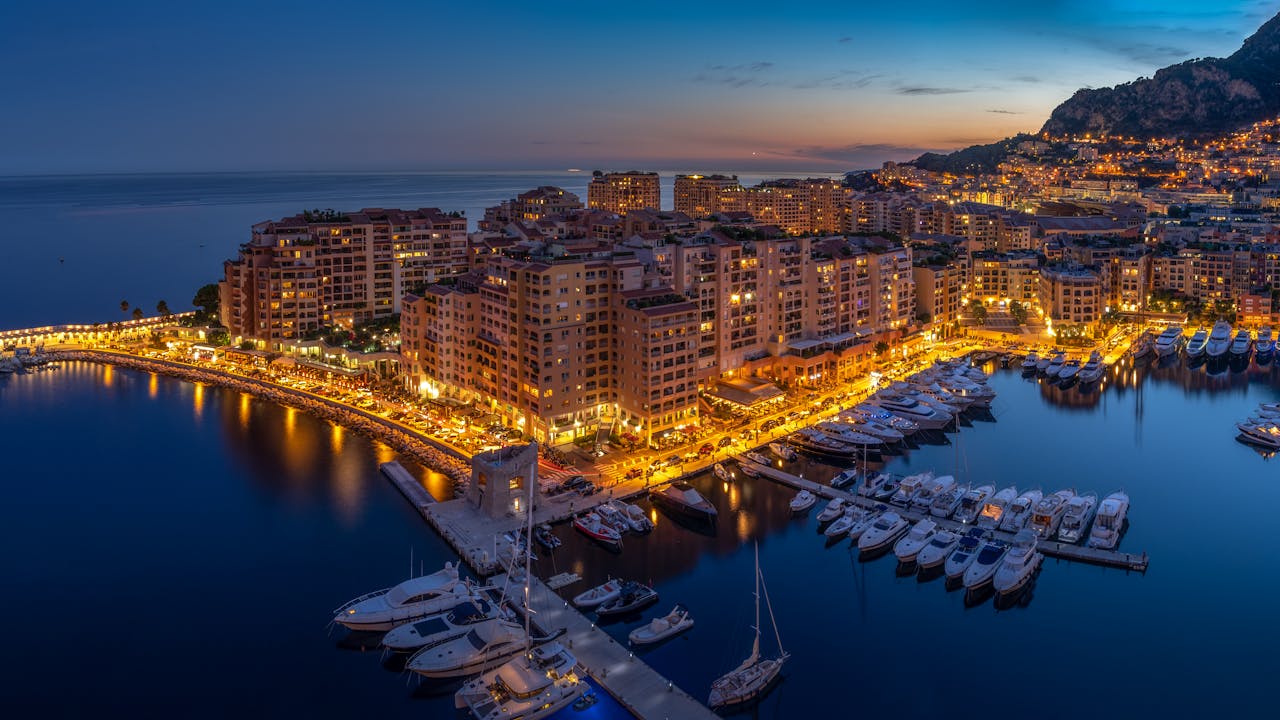 Stunning aerial view of Monaco's harbor at dusk, with city lights glowing and boats docked along the waterfront.